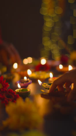 Vertical-Video-Close-Up-Shot-Of-Hands-Lighting-Diya-Oil-Lamps-Celebrating-Festival-Of-Diwali-On-Darkened-Table